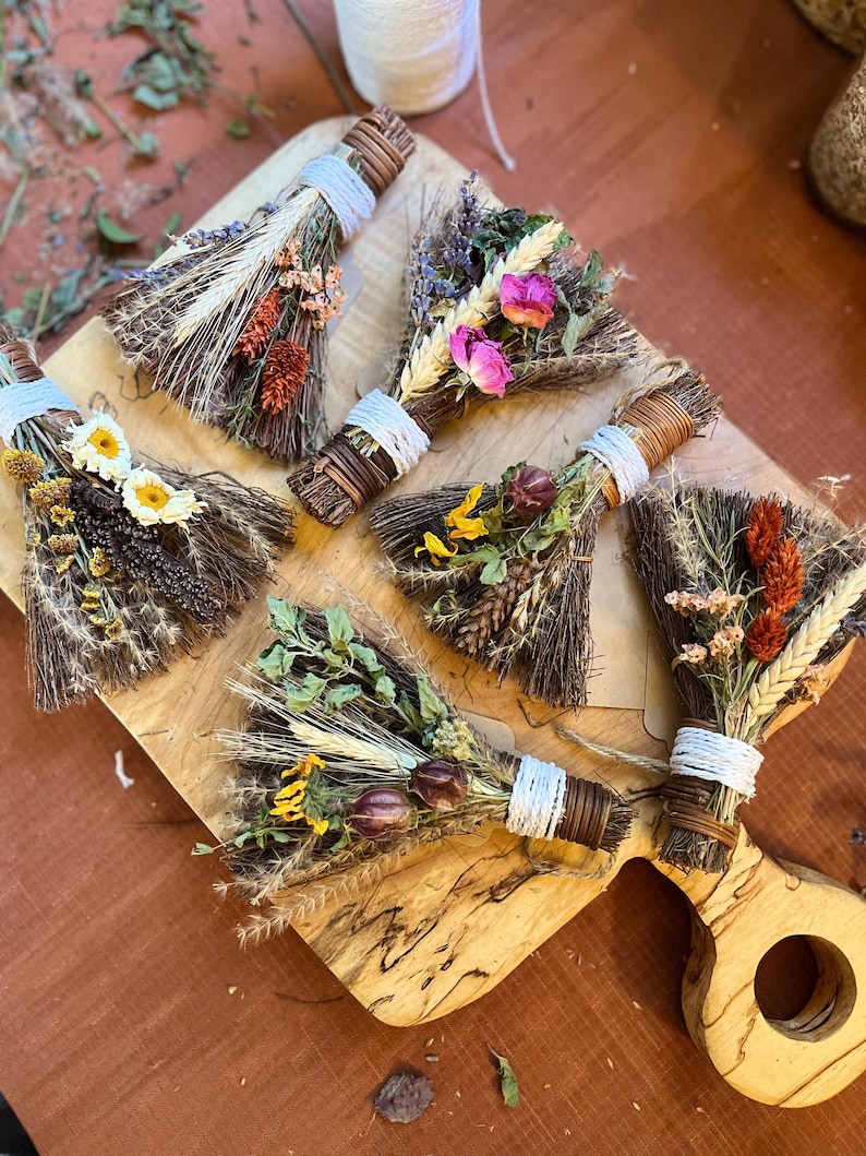 May include: Five small decorative brooms made of dark brown twigs, each tied with white or brown string and adorned with various dried flowers, including pink roses, yellow daisies, orange wheat stalks, and green foliage, arranged on a wooden board.