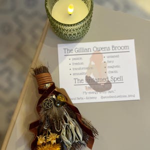 May include: A decorative broom with brown bristles, decorated with feathers, dried yellow flowers, and a brown ribbon. A small card reads "The Gillian Owens Broom" with related words. A green glass candle holder with a lit candle is also present.