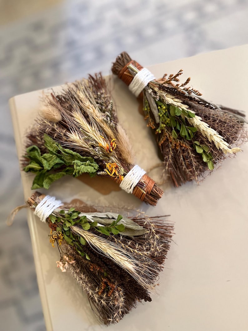 May include: Three decorative bundles of dried herbs and flowers, each tied with white string. The arrangements feature brown twigs, green leaves, wheat stalks, and small orange blossoms, creating a textured, natural appearance for each item.