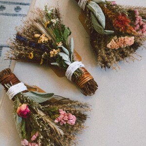 May include: Three small bundles of dried flowers and herbs, each tied with white string and brown twine. The arrangements feature a mix of dried grasses, sage leaves, pink and yellow blossoms, and small sprigs of lavender in a fan-like shape.