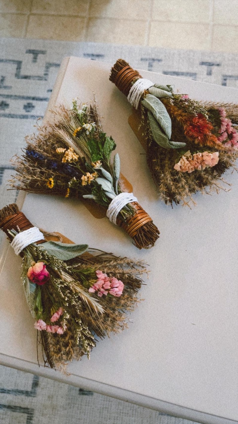 May include: Three small bundles of dried flowers and herbs, each tied with white string and brown twine. The arrangements feature a mix of dried grasses, sage leaves, pink and yellow blossoms, and small sprigs of lavender in a fan-like shape.