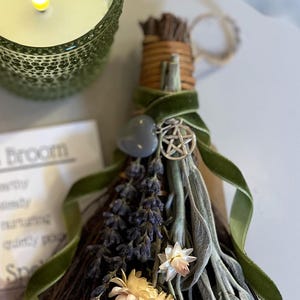 May include: A decorative broom with dried lavender, sage, and flowers, tied with a green velvet ribbon. A silver pentacle charm and a gray heart-shaped stone are attached. A green glass candle holder with a lit candle is in the background.