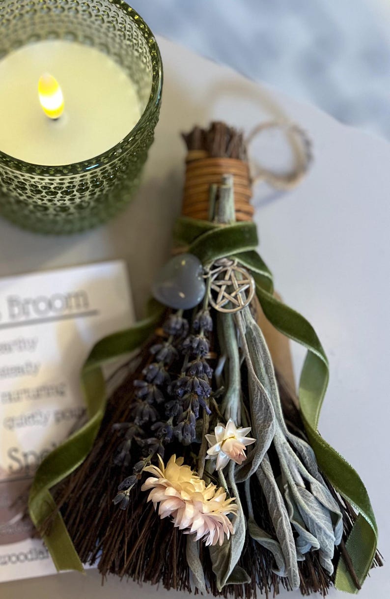 May include: A decorative broom with dried lavender, sage, and flowers, tied with a green velvet ribbon. A silver pentacle charm and a gray heart-shaped stone are attached. A green glass candle holder with a lit candle is in the background.