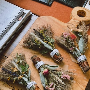 May include: Four small decorative bundles of dried herbs and flowers, each tied with white string and wrapped in thin brown wood strips. The bundles feature various dried botanicals including sage leaves, yellow flowers, and pink blossoms.