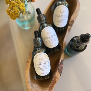 May include: Three glass bottles of Blue Moon face serum in a wooden bowl. The bottles are labeled with the product name and ingredients. The serum is made with a blend of essential oils, including lavender, frankincense, and myrrh.