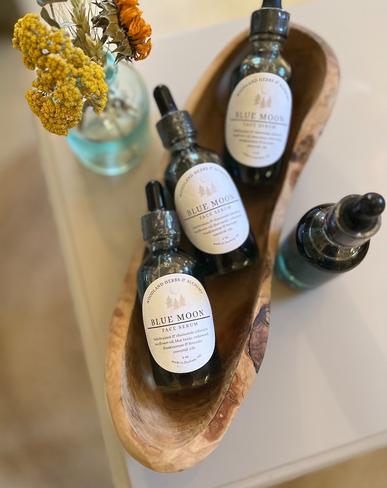 May include: Three glass bottles of Blue Moon face serum in a wooden bowl. The bottles are labeled with the product name and ingredients. The serum is made with a blend of essential oils, including lavender, frankincense, and myrrh.