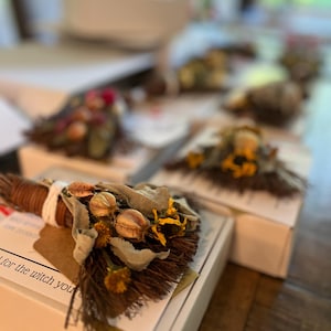 May include: A small decorative broom made of brown twigs, tied with white string, and adorned with dried green leaves, yellow flowers, and round seed pods. The broom is attached to a white box with text that reads for the witch you are.