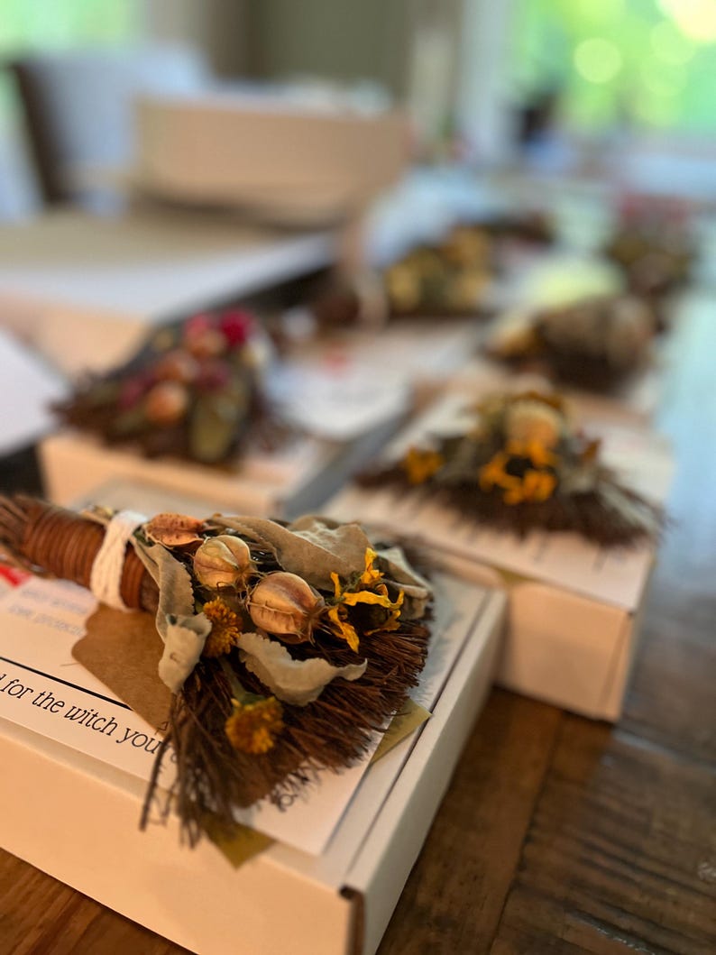 May include: A small decorative broom made of brown twigs, tied with white string, and adorned with dried green leaves, yellow flowers, and round seed pods. The broom is attached to a white box with text that reads for the witch you are.