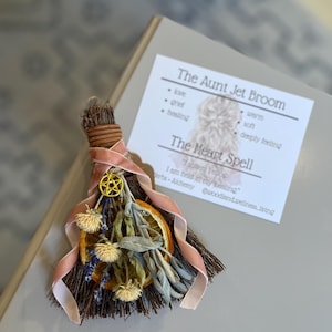 May include: A small decorative broom with dried flowers, an orange slice, and a gold pentacle charm, tied with a pink ribbon. A card next to it reads "The Aunt Jet Broom" and "The Heart Spell". The broom is a unique, handcrafted item.