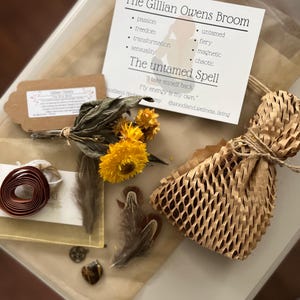 May include: A kit containing a card titled The Gillian Owens Broom with the text The untamed Spell, a bundle of dried yellow flowers, two brown feathers, a small heart-shaped stone, a metal charm, a roll of brown ribbon, and a wrapped item in honeycomb paper.