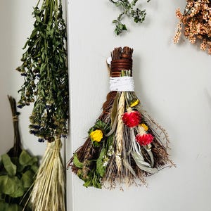 May include: A decorative wall hanging bundle composed of dried twigs, wheat stalks, and vibrant dried flowers including red globe amaranth and yellow blossoms. The stems are tightly bound with brown twine and a thick white cord for hanging display.