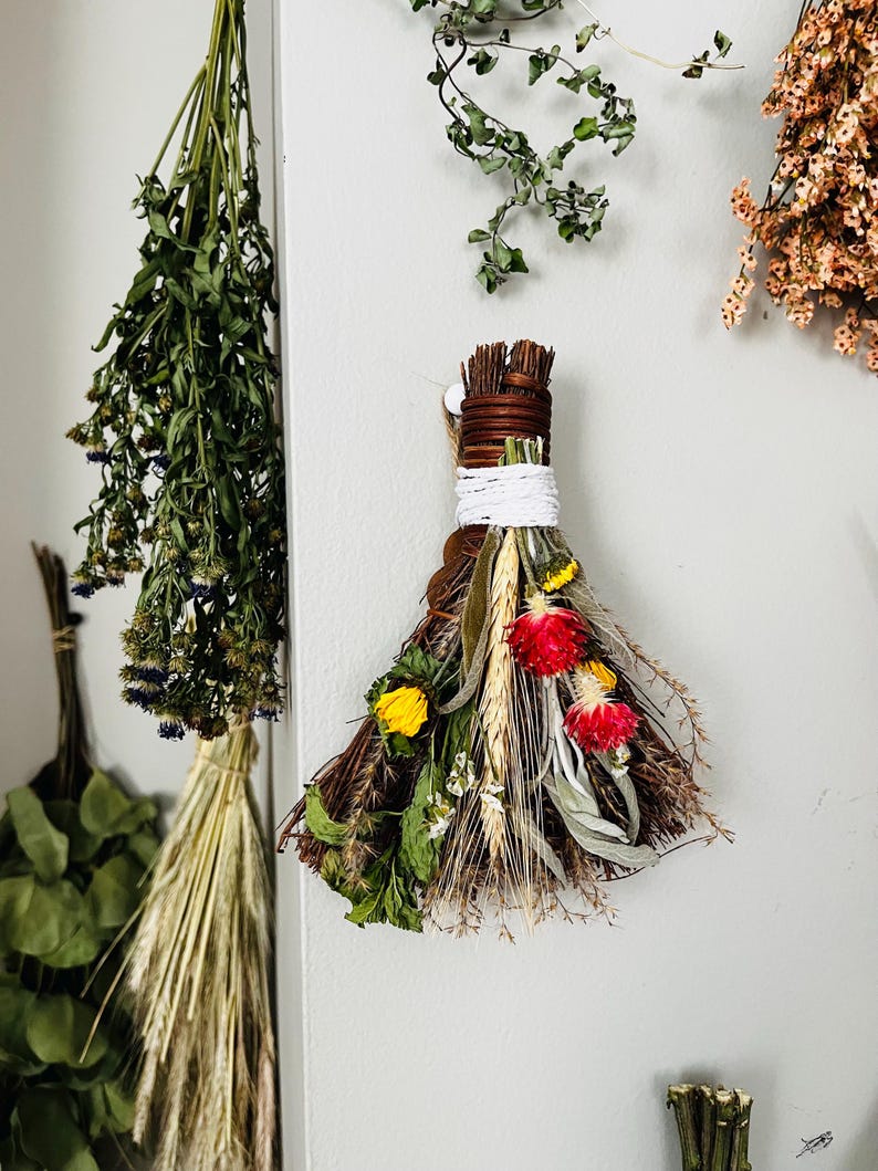 May include: A decorative wall hanging bundle composed of dried twigs, wheat stalks, and vibrant dried flowers including red globe amaranth and yellow blossoms. The stems are tightly bound with brown twine and a thick white cord for hanging display.