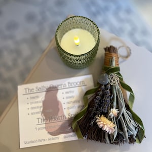 May include: A green glass candle holder with a lit candle next to a small broom decorated with dried flowers, herbs, and a green ribbon. A card with text that reads "The Sally Owens Broom" is also present.