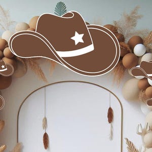 May include: A brown cowboy hat with a white star on the front, surrounded by brown and beige balloons and dried grasses.