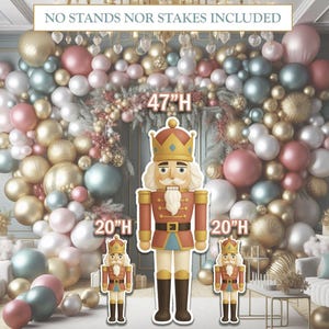 May include: A festive holiday scene featuring a balloon arch in gold, pink, and teal colors. Three nutcracker cutouts are in the foreground, the largest is 47 inches tall, and the smaller ones are 20 inches tall. The text "NO STANDS NOR STAKES INCLUDED" is at the top.