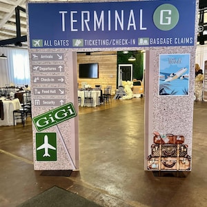 May include: A brown and beige cardboard cutout of a terminal entrance with the text "TERMINAL G" in blue and green. The cutout has a sign with the text "ALL GATES", "TICKETING/CHECK-IN", and "BAGGAGE CLAIMS". There are signs with arrows pointing to "Arrivals", "Departures", "Check-in", "Food Hall", and "Security". There is a sign with the text "GiGi" and a green airplane symbol. The cutout has a stack of brown suitcases with gold accents.