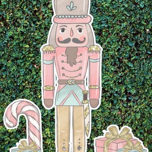 May include: A pink and gold glitter nutcracker with a candy cane and two gift boxes. The nutcracker is wearing a blue and green uniform with a gold belt and a sword. The gift boxes are wrapped in pink and blue paper with gold ribbon.