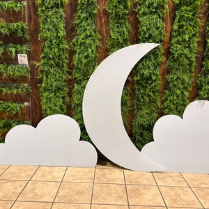 May include: White cardboard cutouts of a crescent moon and two clouds. The moon is large and the clouds are smaller. The cutouts are against a backdrop of green artificial foliage.