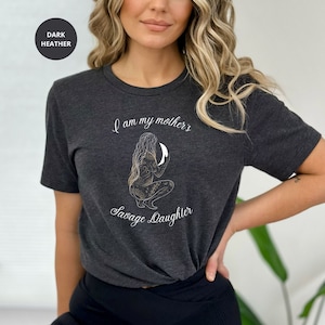 May include: A dark heather grey t-shirt with a graphic of a woman holding a crescent moon. The text "I am my mother's Savage Daughter" is printed on the shirt.