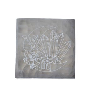 Può includere: Un sottobicchiere quadrato in pietra grigio chiaro con un disegno bianco inciso. Il disegno presenta una luna crescente, cristalli, un fiore e foglie. L'oggetto è decorativo.