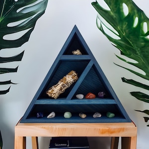 May include: A triangular, navy blue wooden shelf unit with multiple compartments. The shelf holds a bundle of sage, various gemstones, and a piece of pyrite. The shelf is displayed on a wooden table, with large green leaves in the background.