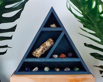 May include: A triangular, navy blue wooden shelf unit with multiple compartments. The shelf holds a bundle of sage, various gemstones, and a piece of pyrite. The shelf is displayed on a wooden table, with large green leaves in the background.