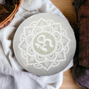 Può includere: Una piastra rotonda in selenite grigio chiaro con un disegno mandala bianco e un simbolo Om al centro. La piastra è appoggiata su un panno bianco, con un cestino di legno e tessuto marrone sullo sfondo.