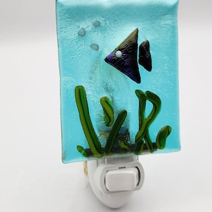 May include: A blue glass night light with a black fish and green seaweed design. The night light plugs into an outlet and has an on/off switch.