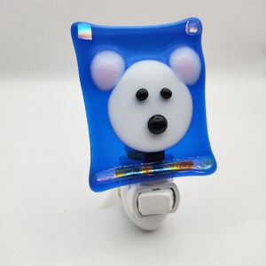 May include: A whimsical night light featuring a polar bear design. The bear's face is white with black eyes and a black nose, set against a vibrant blue background. The night light has a white base with an on/off switch.