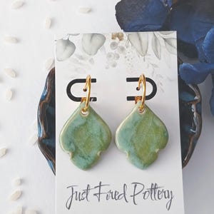 May include: A pair of green ceramic earrings with gold-colored hoops. The earrings are in a unique shape and are displayed on a white card with the text "Just Fired Pottery". The background includes blue decorative elements.