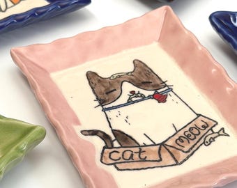 May include: Handmade ceramic trays with a whimsical cat design. Each tray features a unique color and a cartoon cat illustration with the word "cat" and "meow". The trays are rectangular with wavy edges.