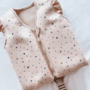 May include: A pink sleeveless life vest with a zip closure and a star pattern in various colours. The vest has a strap with a buckle.