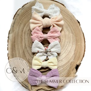 May include: A set of six hair bows in various colors and patterns, including pink, white, yellow, and floral print. The bows are arranged on a wooden surface and are part of a collection called "The Summer Collection".