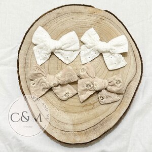 May include: Four handmade hair bows, two white and two beige, with a delicate lace pattern. The bows are displayed on a wooden surface with a logo that reads "C&M estd 2022".