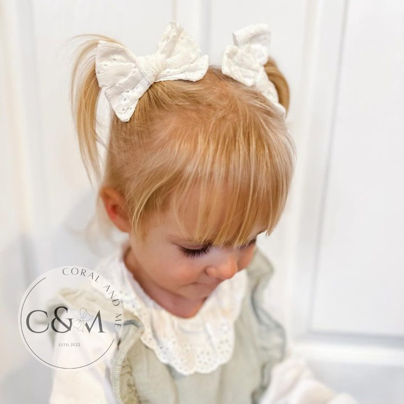 May include: Two white cotton eyelet bows with a white elastic band. The bows are tied in a simple knot and are worn in a little girl's hair.