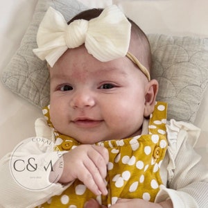 May include: A baby wearing a yellow and white polka dot jumpsuit with a white knotted bow headband. The headband is made of a soft, textured fabric. The baby is smiling and looking at the camera.