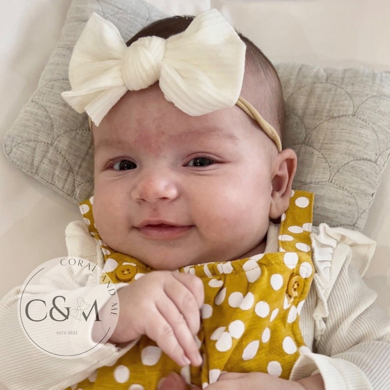 May include: A baby wearing a yellow and white polka dot jumpsuit with a white knotted bow headband. The headband is made of a soft, textured fabric. The baby is smiling and looking at the camera.