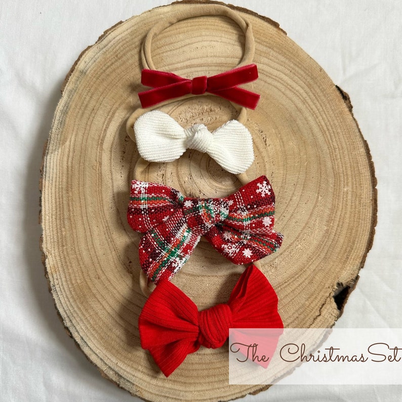 May include: Set of four Christmas-themed hair bows. The bows are red, white, and plaid with a snowflake pattern. The bows are on a wooden surface. The text "The Christmas Set" is visible at the bottom of the image.