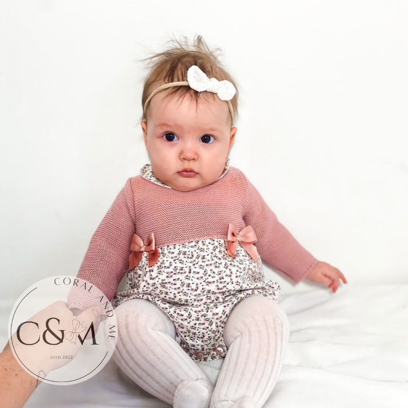 May include: A baby wearing a pink and white floral knitted onesie with a white bow headband. The onesie has a pink knitted top and a white floral bottom. The baby is sitting on a white surface.
