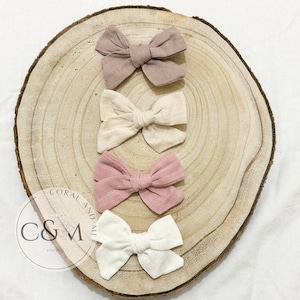 May include: Four fabric hair bows in different colors: beige, light pink, dark pink, and white. The bows are arranged on a wooden surface.