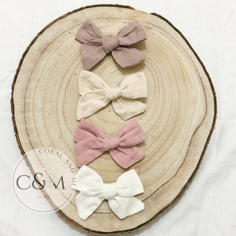 May include: Four fabric hair bows in different colors: beige, light pink, dark pink, and white. The bows are arranged on a wooden surface.