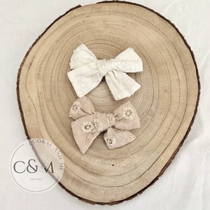 May include: Two fabric hair bows on a wooden surface. The top bow is white with a textured pattern. The bottom bow is beige with a floral lace pattern. The bows are both tied in a bow shape.