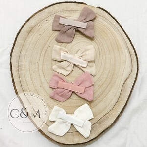 May include: Four fabric hair bows in different colors. The bows are arranged in a circle on a wooden surface. The bows are made of a soft, textured fabric and have a simple, elegant design. The colors of the bows are beige, light pink, dark pink, and white.