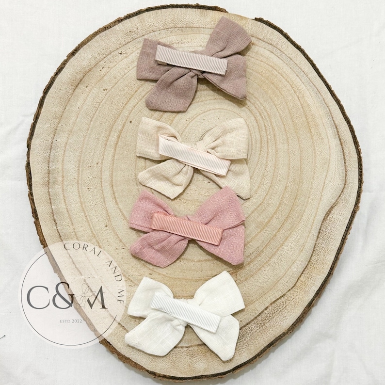 May include: Four fabric hair bows in different colors. The bows are arranged in a circle on a wooden surface. The bows are made of a soft, textured fabric and have a simple, elegant design. The colors of the bows are beige, light pink, dark pink, and white.