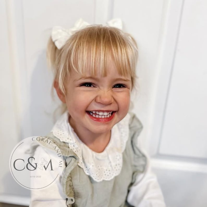 May include: A young girl with blonde hair and a white bow in her hair smiles at the camera. She is wearing a green corduroy dress with a white lace collar and a white long-sleeved shirt underneath. The dress has ruffles on the shoulders and the collar. The girl is standing in front of a white door. The image includes a logo for "Coral and Me" with the text "C&M estd 2022".