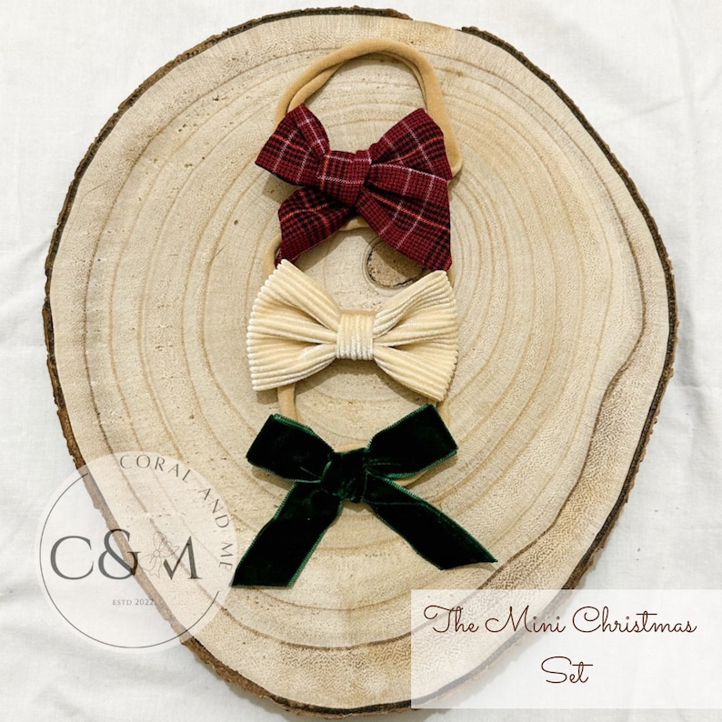 May include: Set of three mini Christmas hair bows. The bows are red plaid, cream velvet, and green velvet. The bows are on a wooden background with the text "The Mini Christmas Set" and a logo that says "Coral and Me".