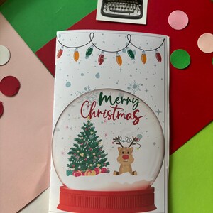 May include: A white card with a red and white snow globe design. The snow globe features a green Christmas tree, a red reindeer, and the text "Merry Christmas".