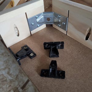May include: A metal corner bracket securing two light-colored wooden boards together with screws, accompanied by three separate black plastic mounting components placed on a flat surface, each featuring pre-drilled holes for installation.