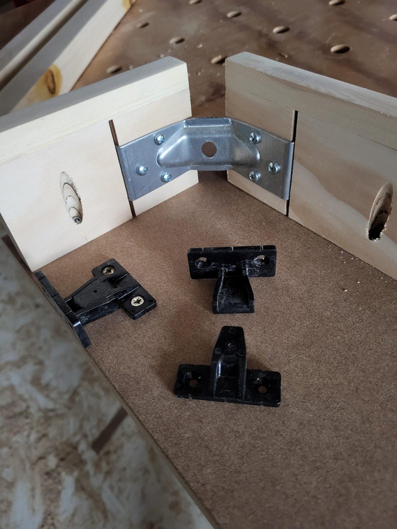 May include: A metal corner bracket securing two light-colored wooden boards together with screws, accompanied by three separate black plastic mounting components placed on a flat surface, each featuring pre-drilled holes for installation.