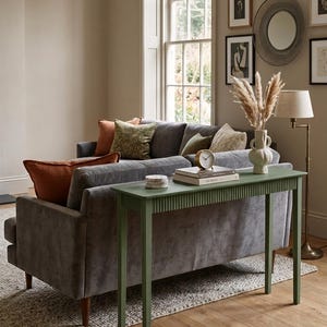 May include: A living room interior with a grey sofa and various cushions in orange, green, and cream. A green console table with a ribbed design supports books, a clock, and coasters. A vase with dried flowers and a floor lamp add to the room's ambiance.